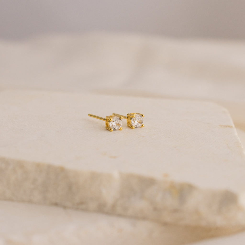 The Booker Large Diamond Studs are minimalist gold earrings with clear, square-cut gemstones, elegantly displayed on a light cream-colored stone surface.