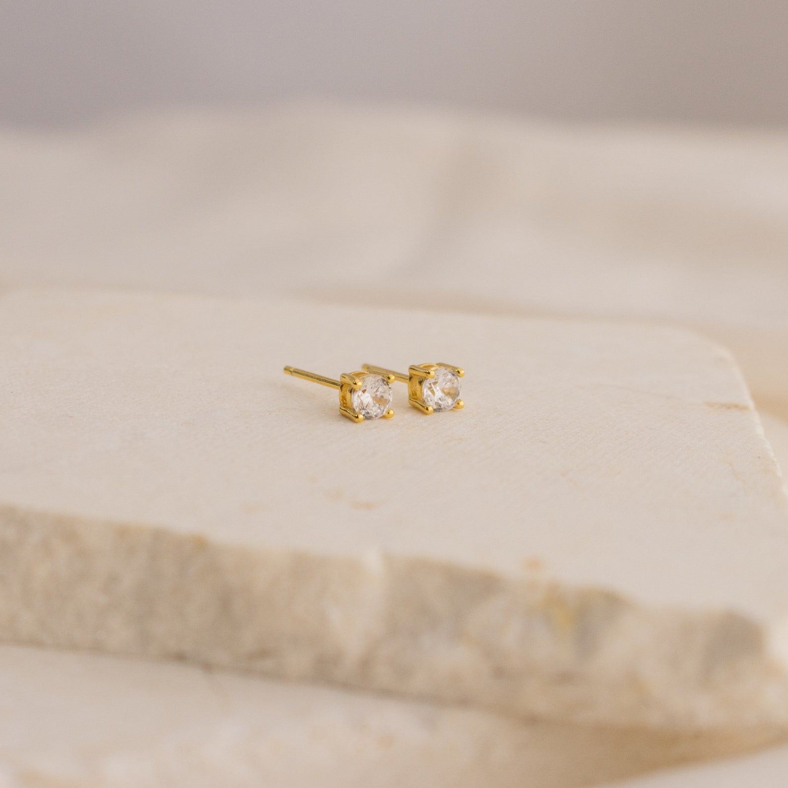 The Booker Large Diamond Studs are minimalist gold earrings with clear, square-cut gemstones, elegantly displayed on a light cream-colored stone surface.