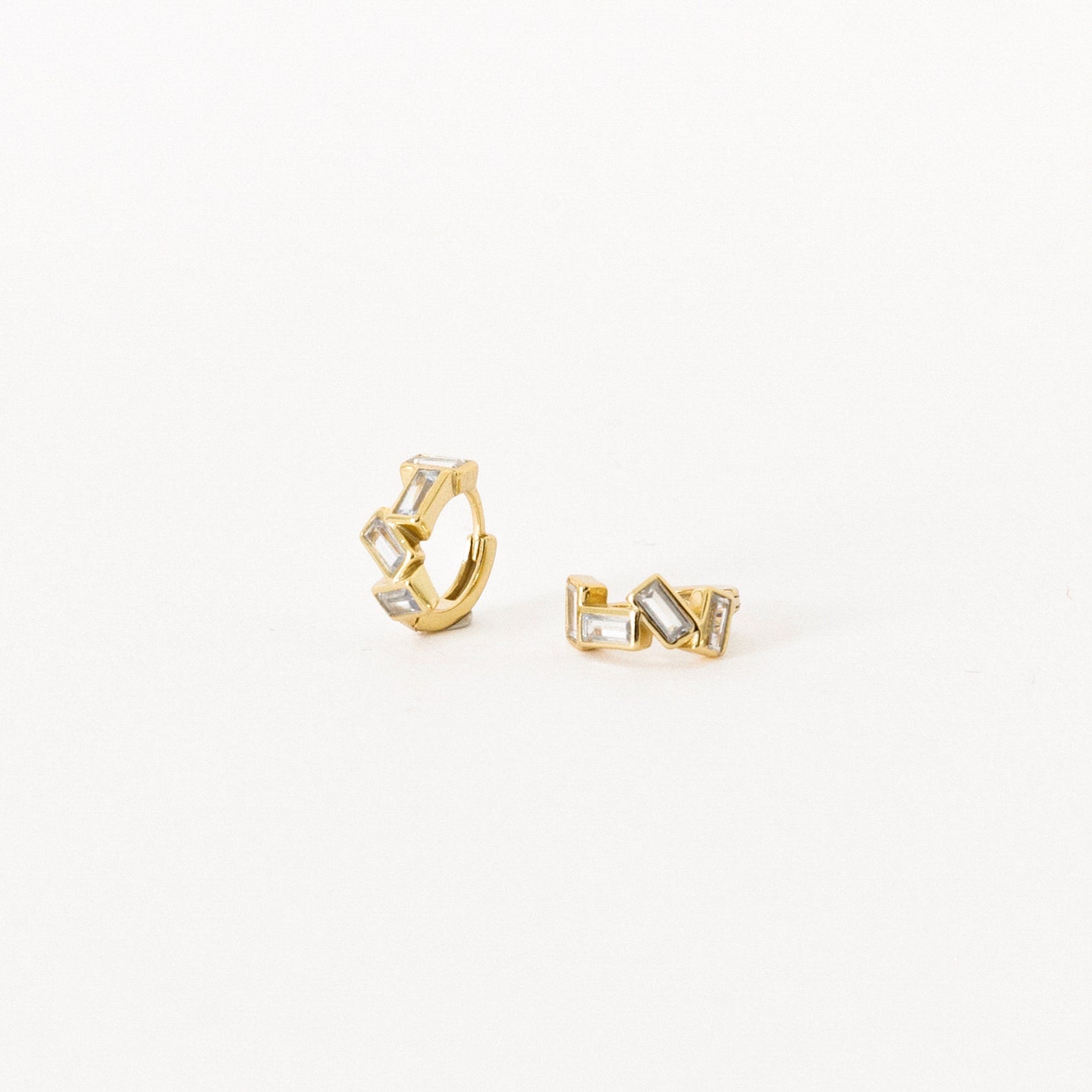 The Glacier Baguette Huggies feature gold hoops with geometric shapes and rectangular clear stones for minimalistic sparkle, elegantly shown on a white background.