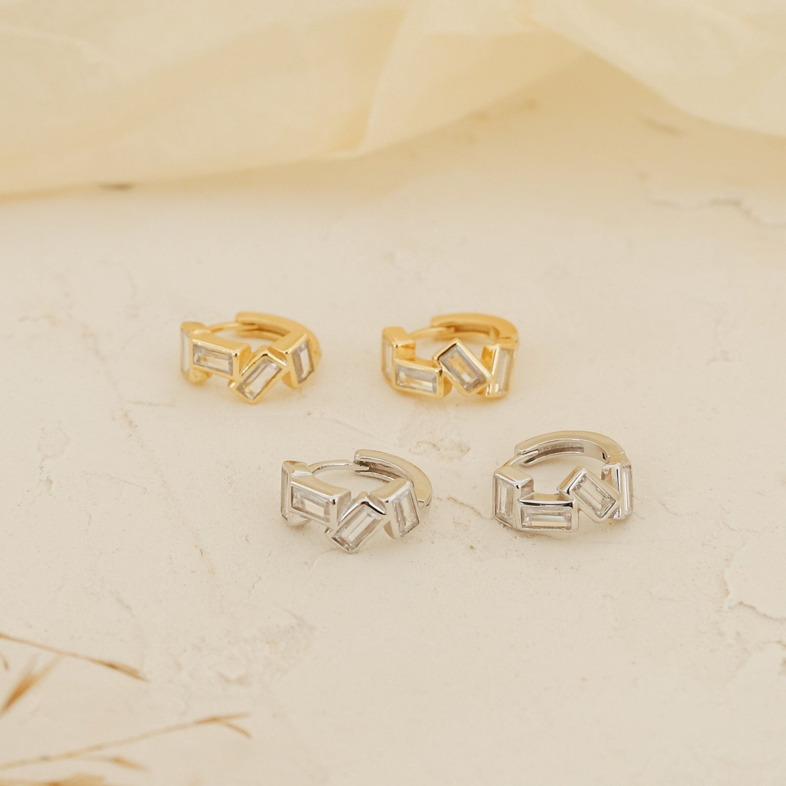 The Glacier Baguette Huggies, featuring two gold and two silver ear cuffs with a minimalistic sparkle, are displayed on a cream-colored textured surface.