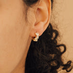 A close-up shows a woman's ear wearing Glacier Baguette Huggies, set against her curly dark hair and highlighting their minimal, sparkling design.