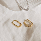 Two chic Indiana Beaded Hoops with beaded edges rest on a white textured fabric beside a clear ribbed glass.
