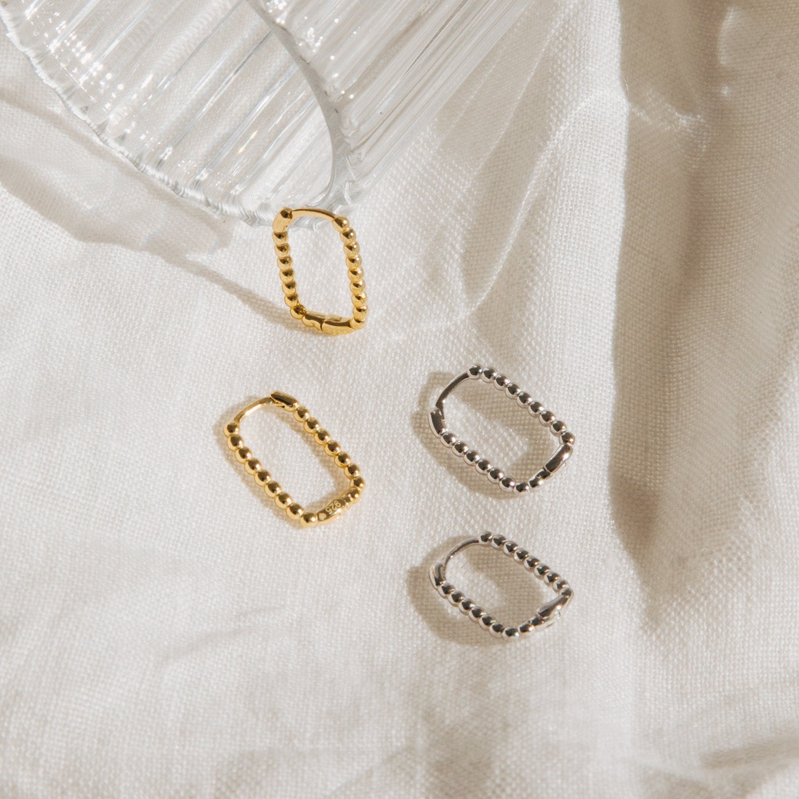 Four stylish Indiana Beaded Hoops—two gold and two silver—are displayed on white fabric by a glass cup, bringing a modern accent to your accessory collection.