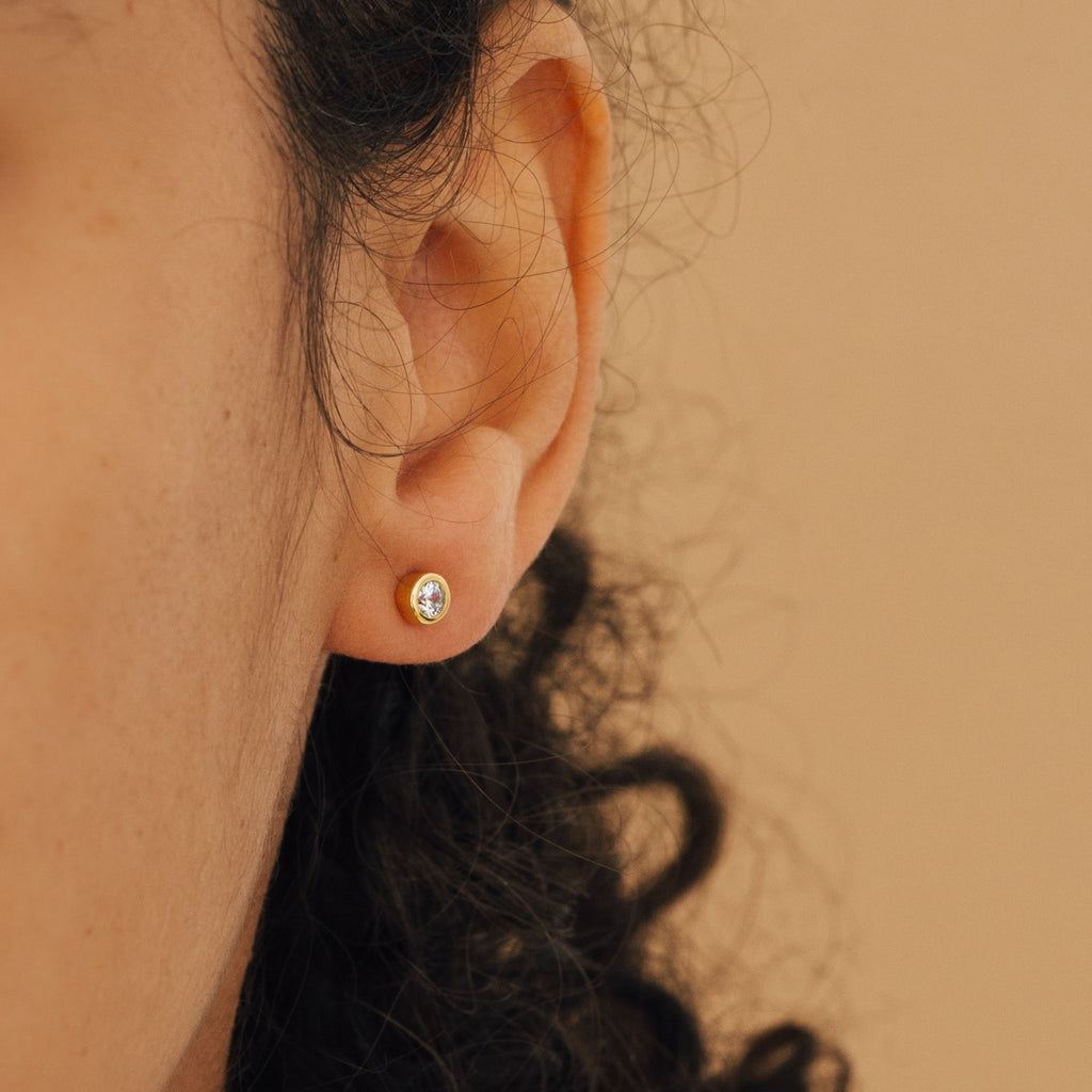 Close-up of a person's ear wearing the Jane Diamond Studs—gold earrings with bezel set diamonds—shown against a beige background.
