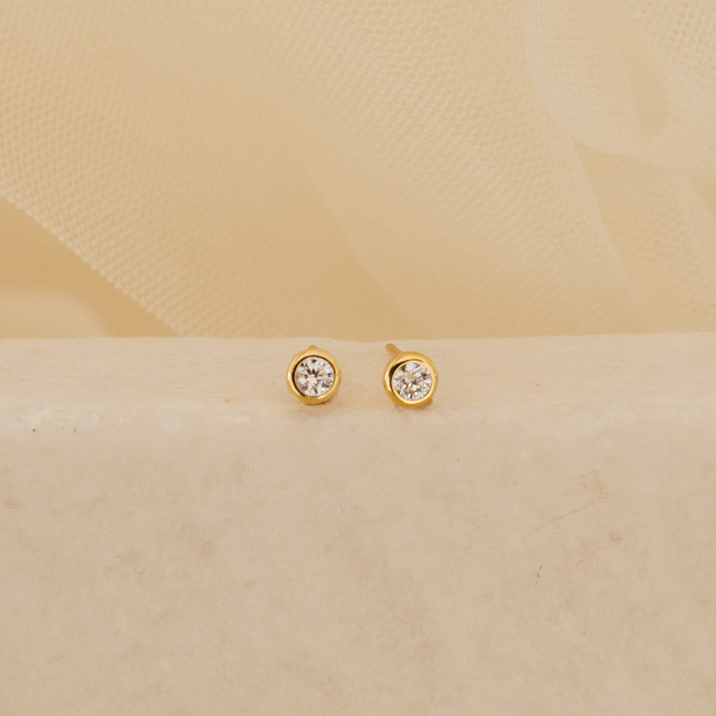 The Jane Diamond Studs are gold earrings with round brilliant diamonds, shown on a light beige surface against a soft background.