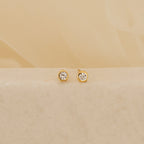 The Jane Diamond Studs are gold earrings with round brilliant diamonds, shown on a light beige surface against a soft background.