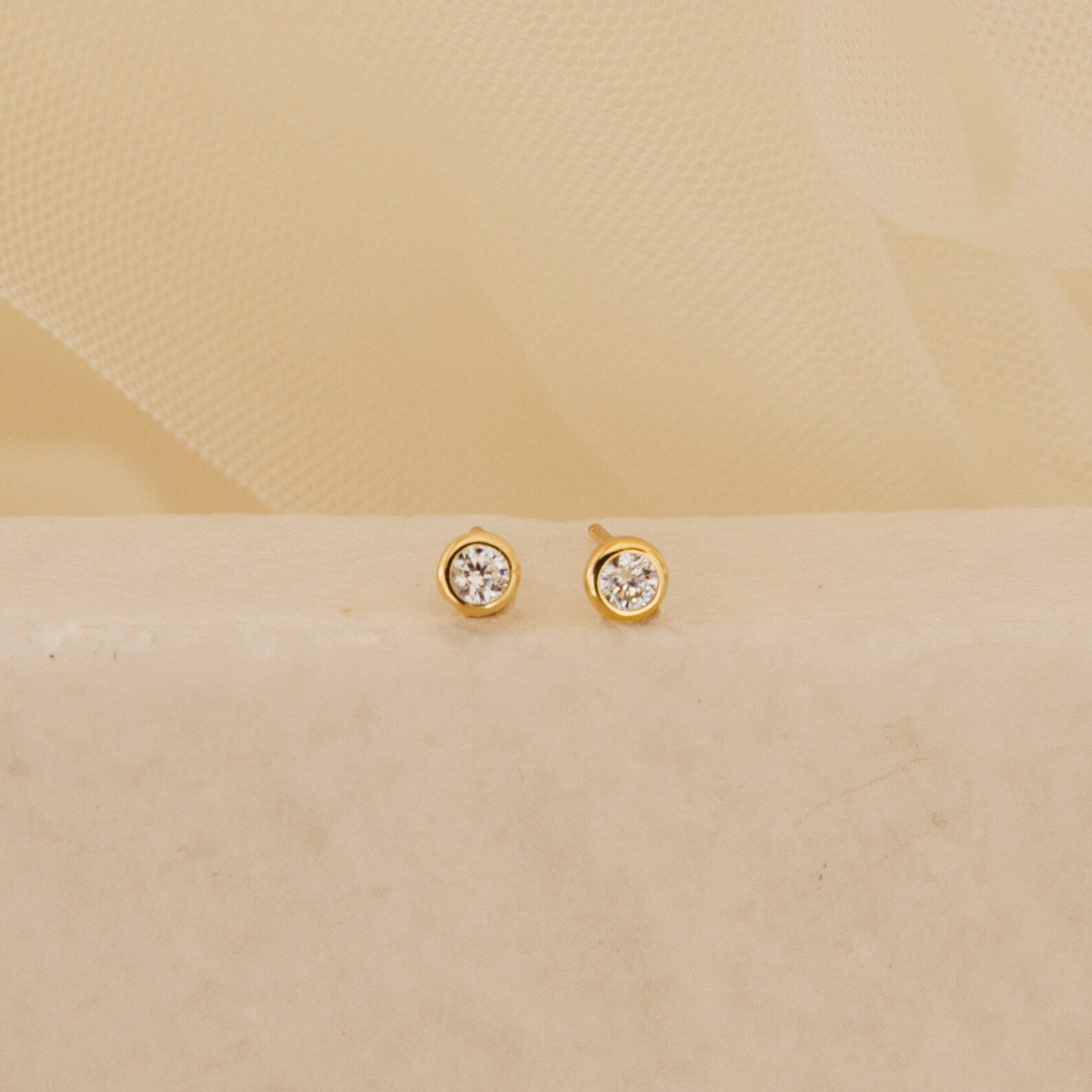 The Jane Diamond Studs are gold earrings with round brilliant diamonds, shown on a light beige surface against a soft background.
