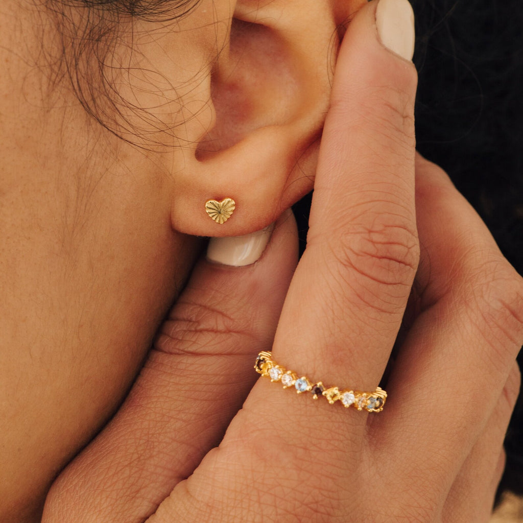 Close-up of a gold heart-shaped stud earring and the Rainbow Crystal Ring, a vintage-inspired band with pastel-toned gemstones, shown on a person's hand and ear.