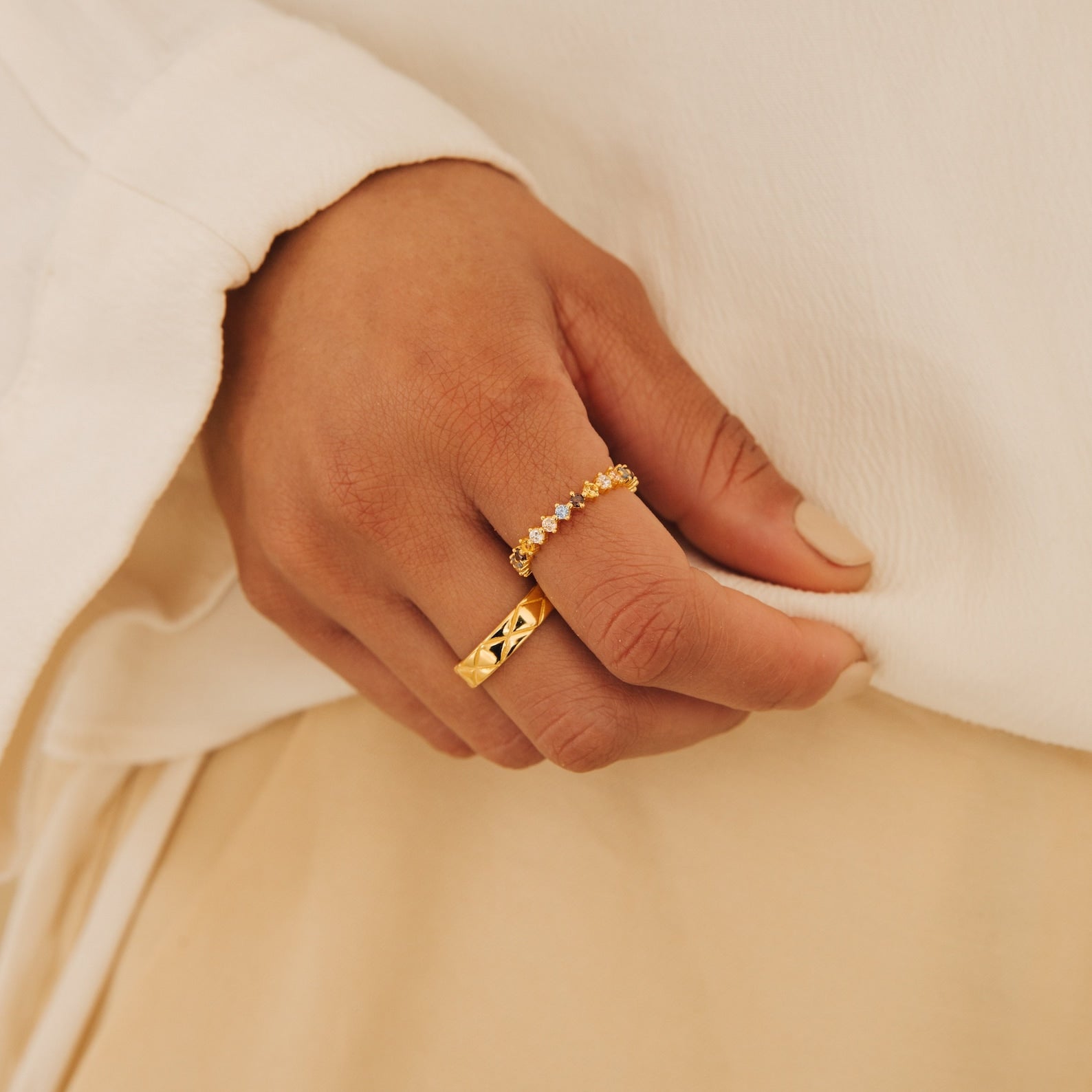 A hand wearing the Rainbow Crystal Ring, featuring pastel toned gemstones, rests on light beige fabric; nails painted nude.