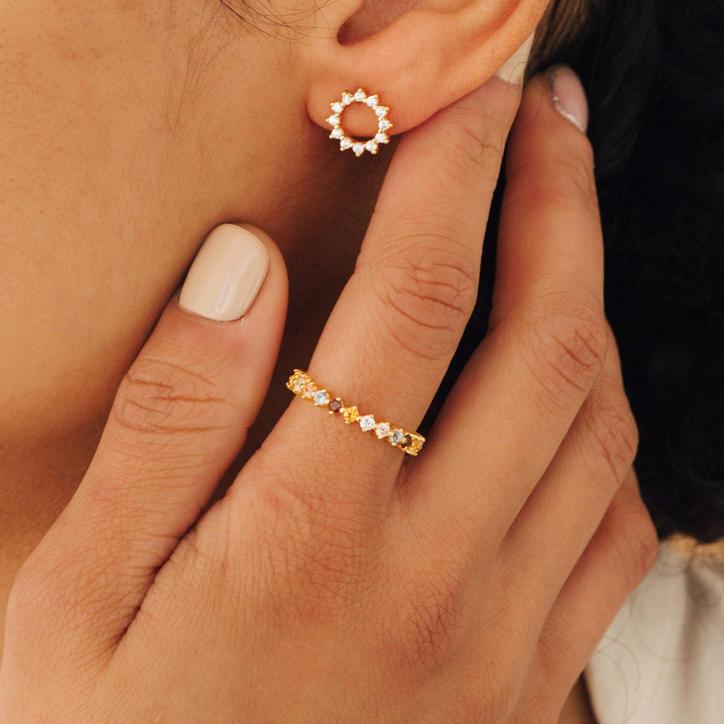 A hand with light nude nails touches an ear wearing a gold and diamond stud; the Rainbow Crystal Ring, vintage-inspired, adorns the finger alongside a matching gold band.