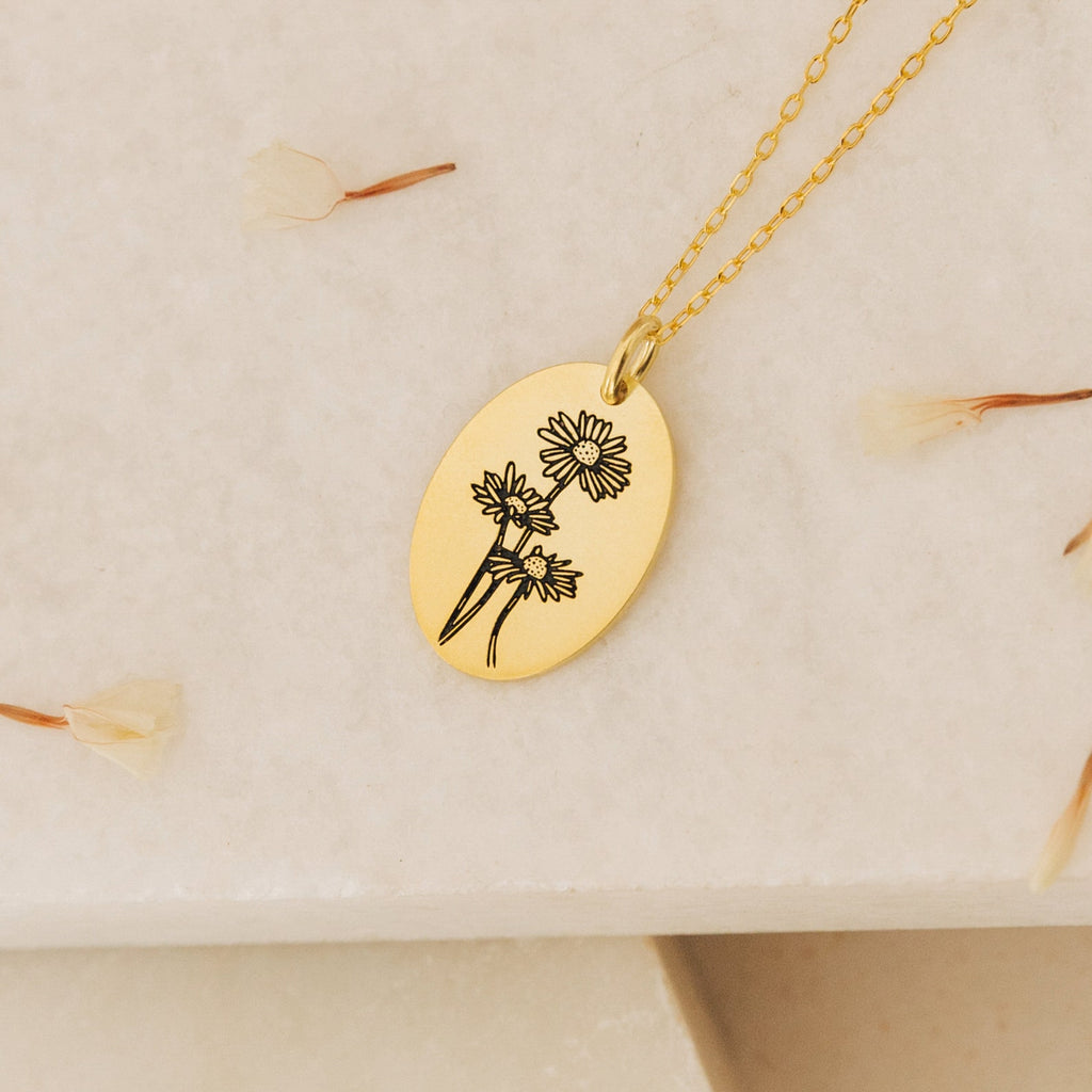 The Oval Flower Necklace features an engraved gold oval pendant with three flowers, displayed on a light textured surface with dried petals—an elegant birth flower necklace, perfect for unique flower jewelry lovers.