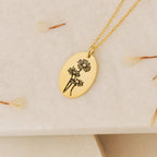 The Oval Flower Necklace features an engraved gold oval pendant with three flowers, displayed on a light textured surface with dried petals—an elegant birth flower necklace, perfect for unique flower jewelry lovers.
