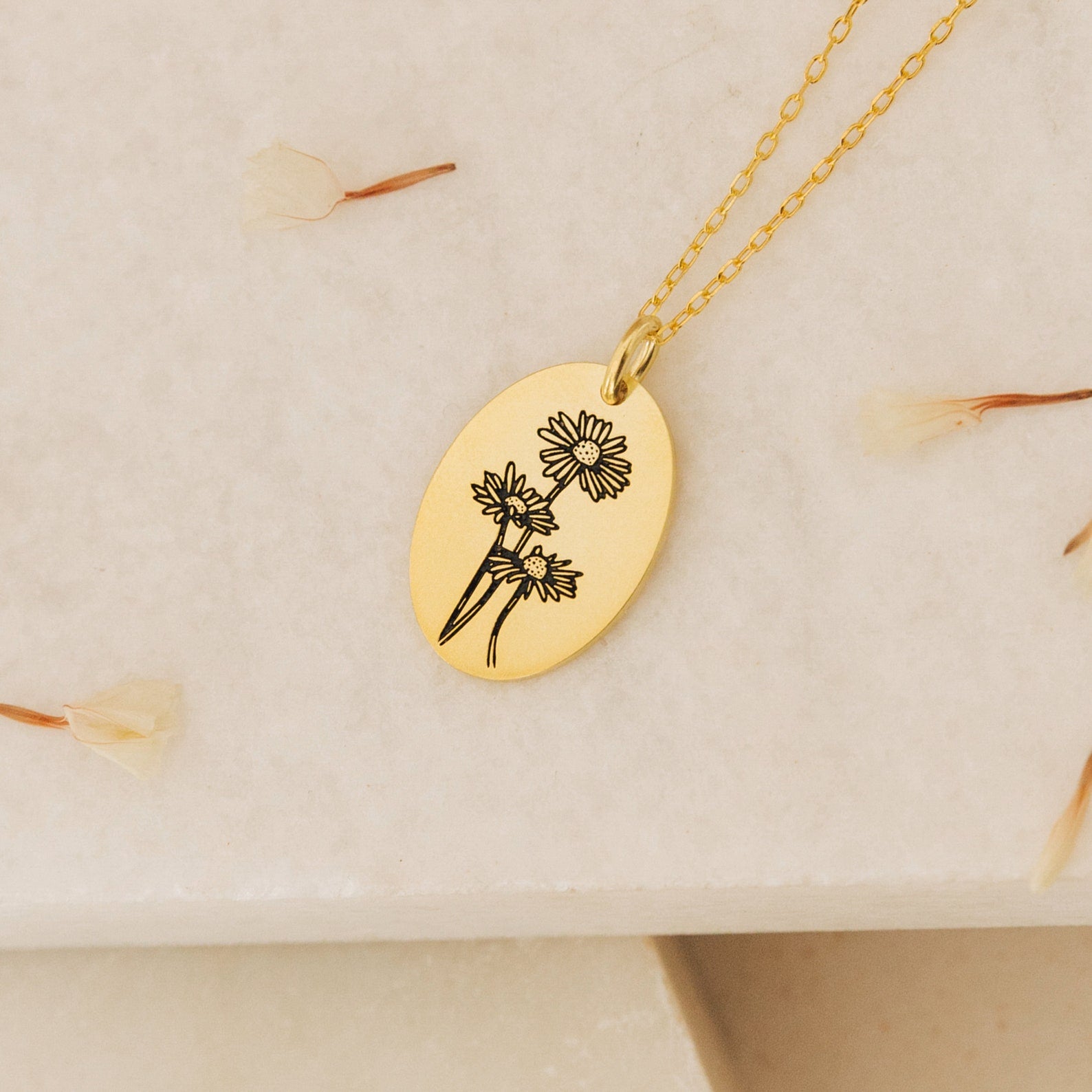 The Oval Flower Necklace features an engraved gold oval pendant with three flowers, displayed on a light textured surface with dried petals—an elegant birth flower necklace, perfect for unique flower jewelry lovers.