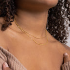 A woman with curly hair in a beige off-shoulder top wears the elegant Monet Duo Chain necklace paired with another delicate gold necklace.