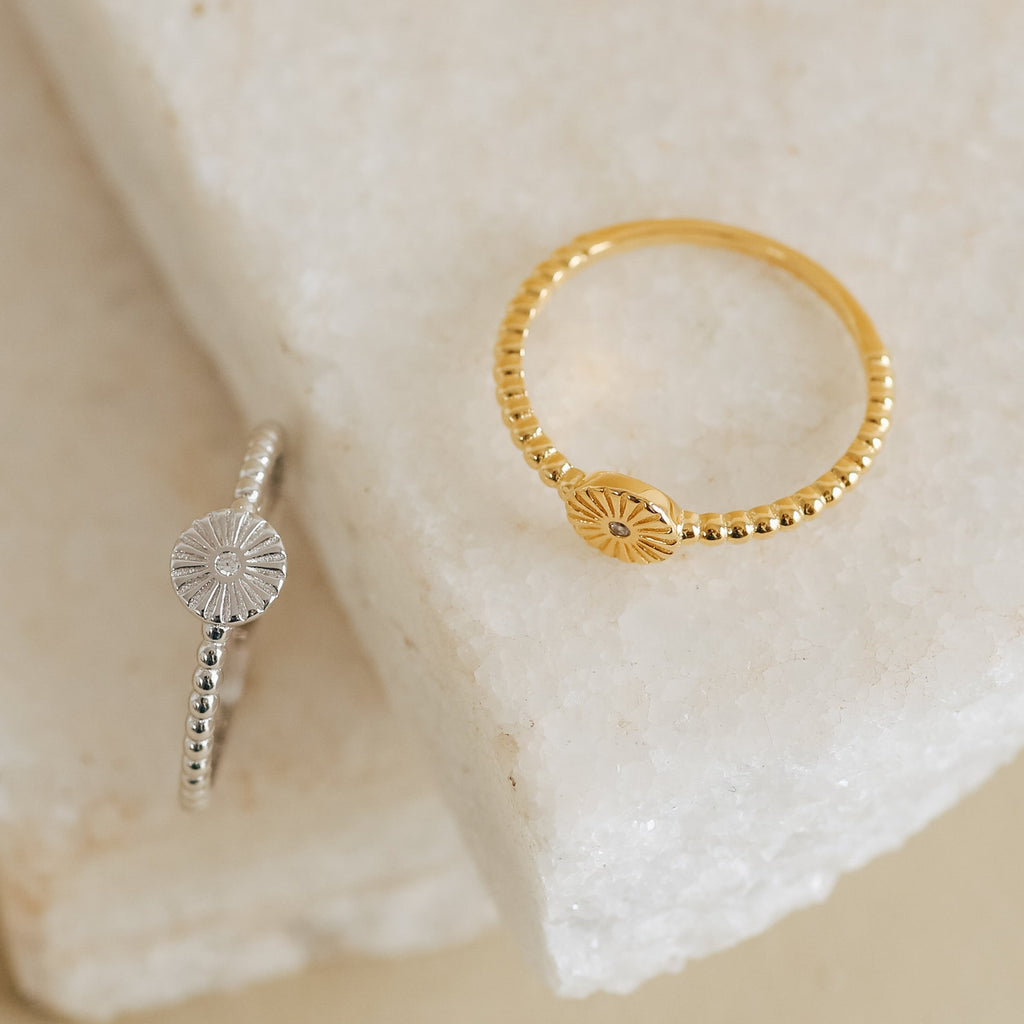 The Eden Sun Ring, featuring a radiant sunburst design and beaded band in silver, exudes boho-chic charm as it rests stylishly on a white stone surface.