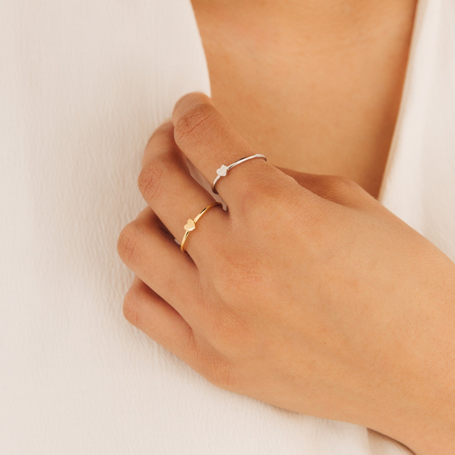 A hand wears two Nala Tiny Heart Rings—one gold, one silver—with petite heart designs, set against a white fabric background.