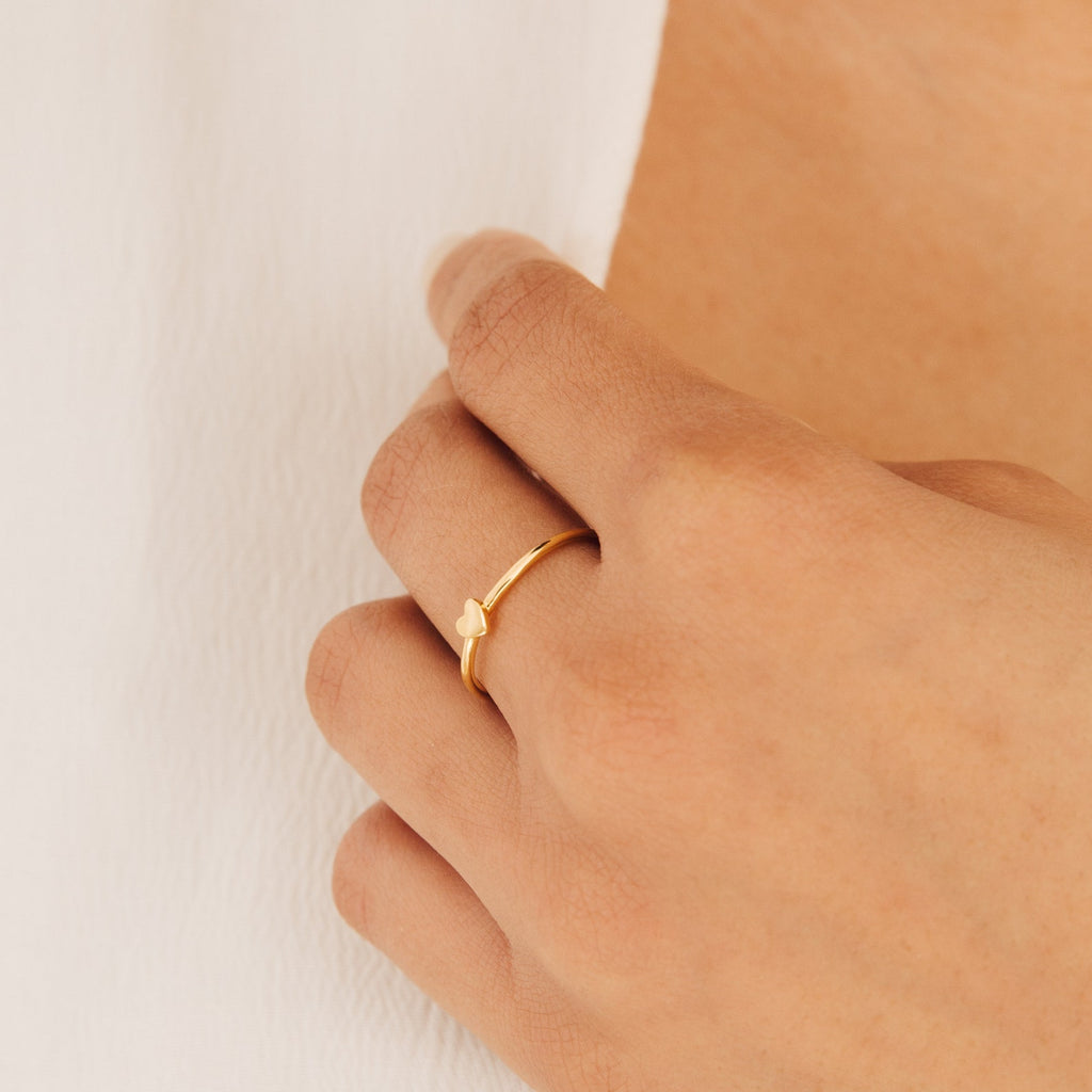 A hand wears the Nala Tiny Heart Ring, featuring a delicate heart-shaped detail—perfect as a promise ring—resting gently on white fabric.