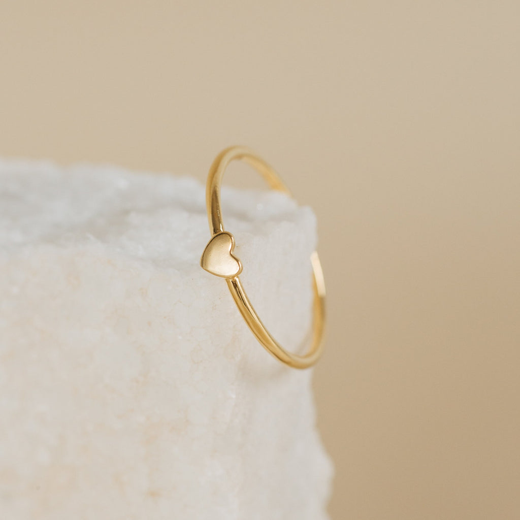 The Nala Tiny Heart Ring, featuring a small heart detail, rests on a textured white surface with a beige background.