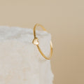 The Nala Tiny Heart Ring, featuring a small heart detail, rests on a textured white surface with a beige background.
