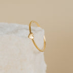The Nala Tiny Heart Ring, featuring a small heart detail, rests on a textured white surface with a beige background.