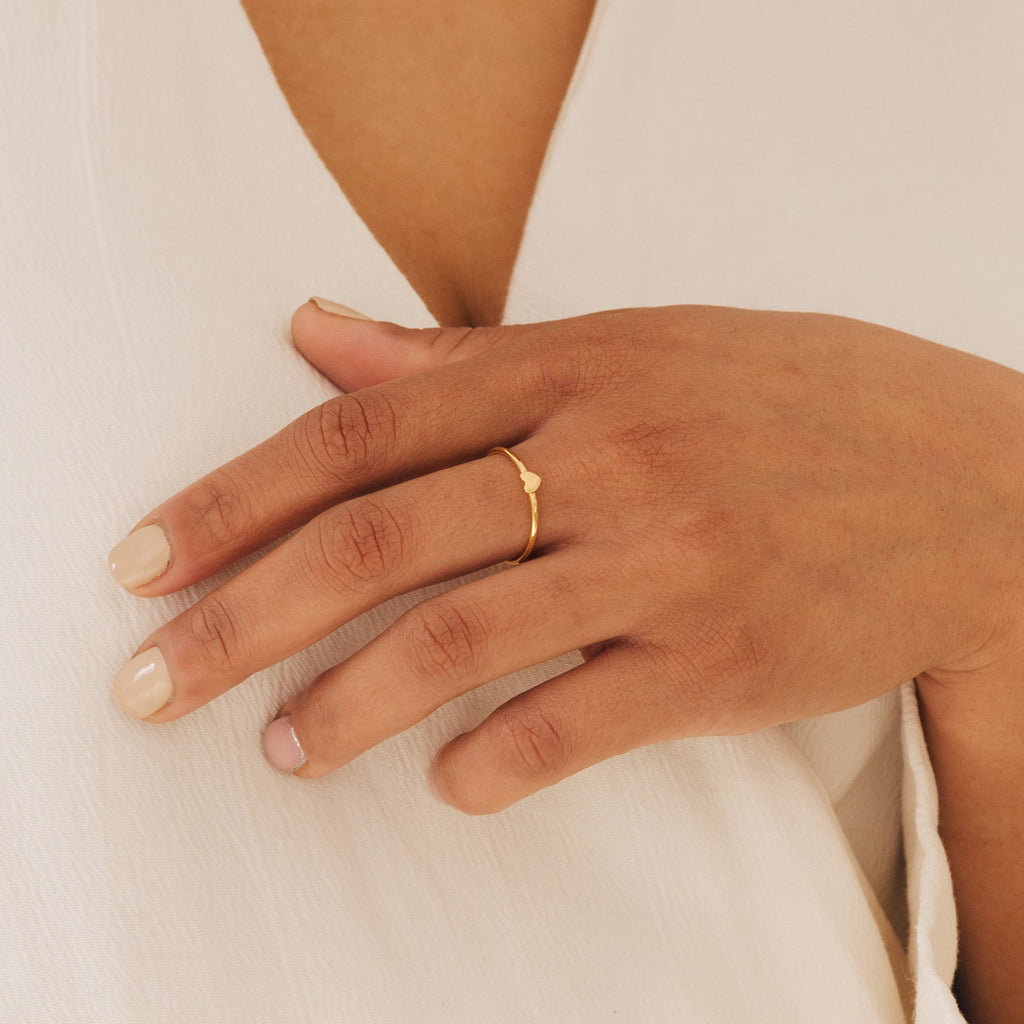 A hand with light nail polish wears the Nala Tiny Heart Ring, delicately resting on a cream-colored v-neck fabric.