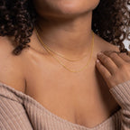 Close-up of a woman wearing two layered gold necklaces: a sleek, flat snake chain and a faceted bead-style chain, styled with a ribbed off-the-shoulder top.