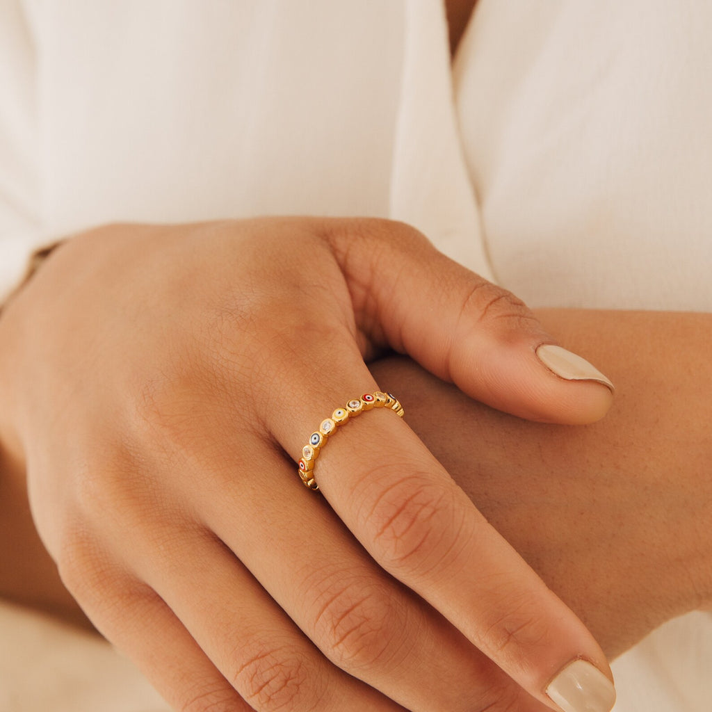 A hand with nude nails wears the Rainbow Evil Eye Ring, a gold piece adorned with small clear stones, resting on cream-colored fabric.