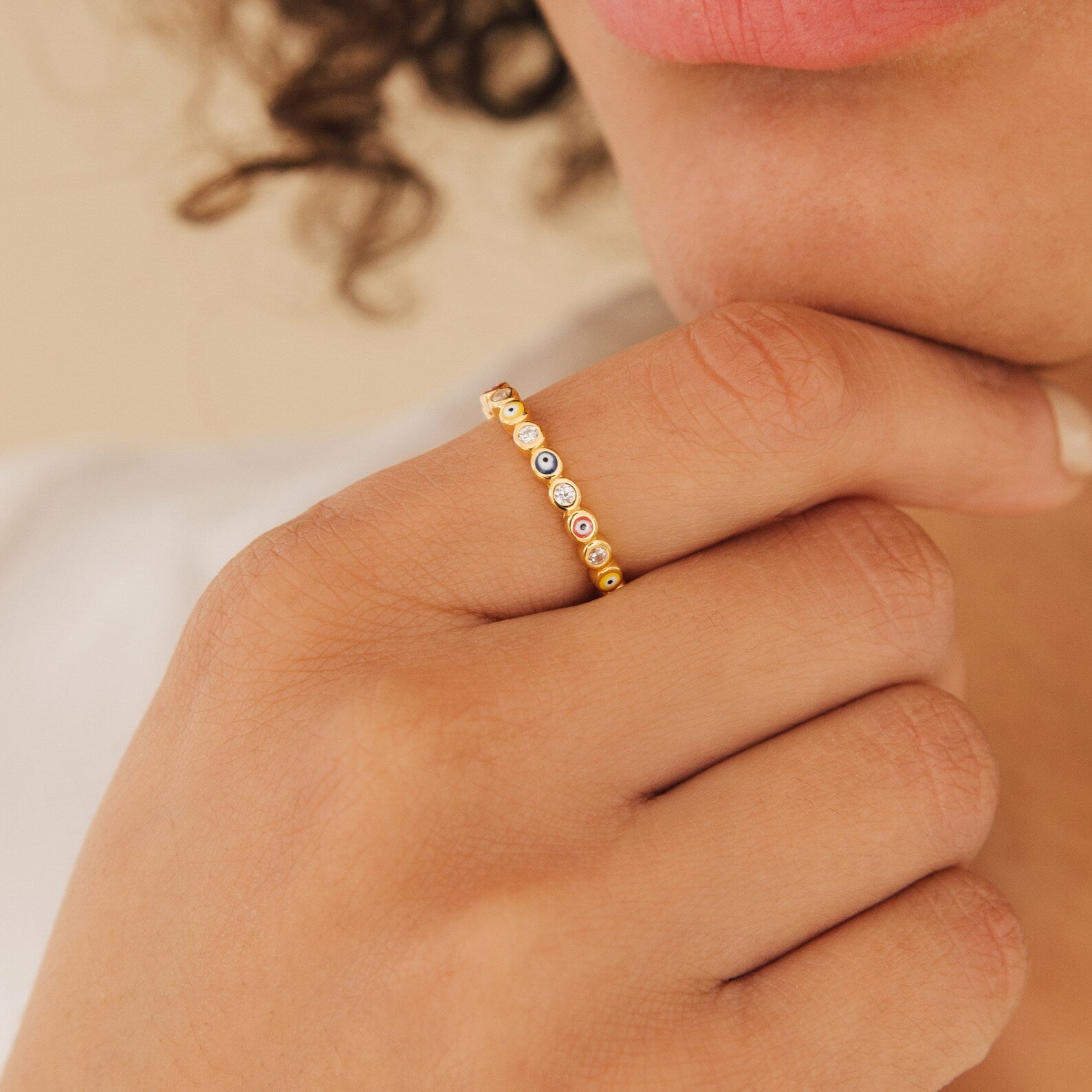 A hand with curly hair is adorned with the Rainbow Evil Eye Ring, a gold band set with small colorful stones, held close to the chin.