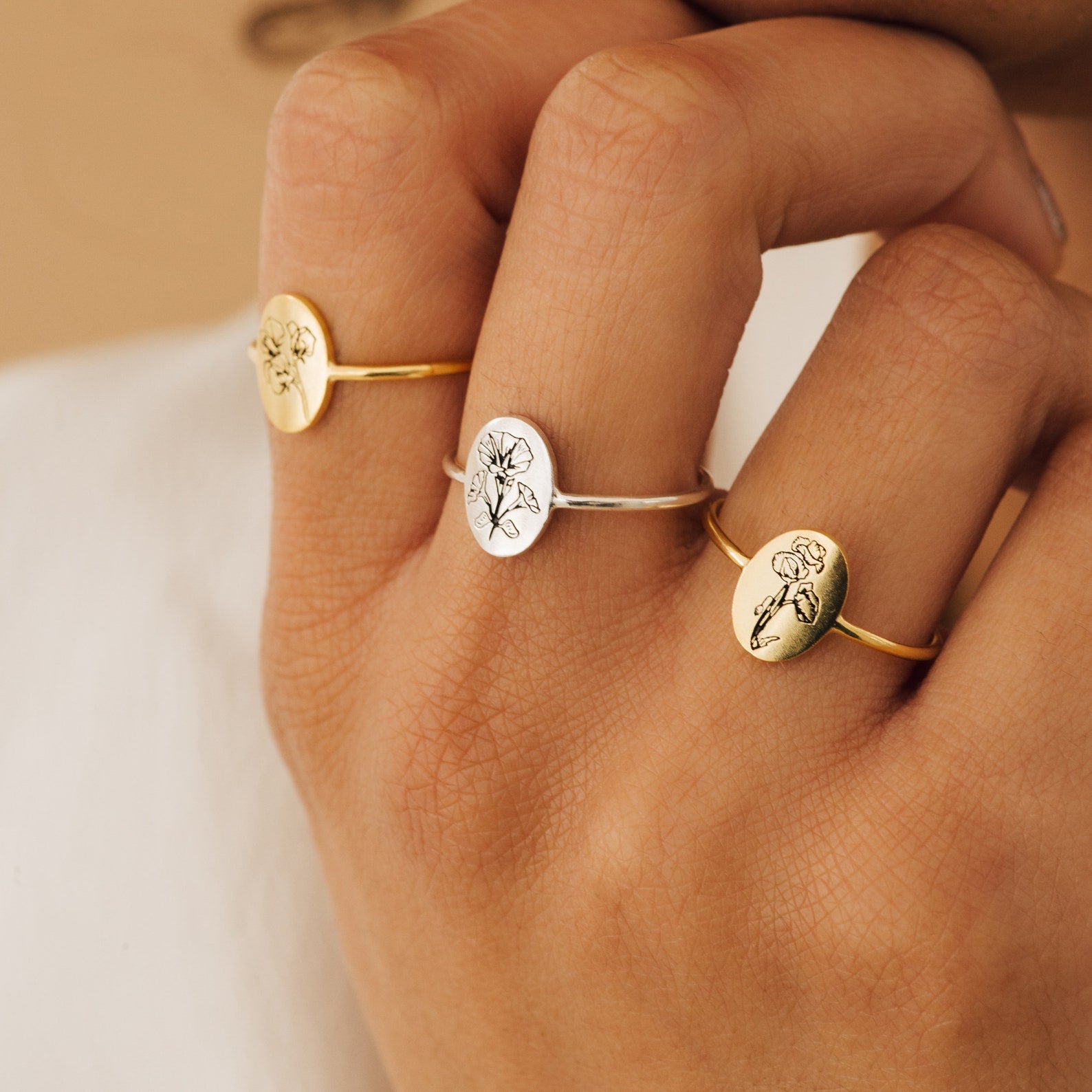 Three engraved Flower Oval Rings—gold and silver—worn on a person's fingers, close-up view of hand. Each delicate ring is perfect for those who love personalized floral jewelry.