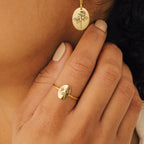 A woman wears the Flower Oval Ring—a gold birth flower ring with rose engravings—on her hand, along with matching earrings; her nails are painted a nude color.