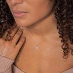 A woman wears the Athens Diamond Necklace, featuring a delicate gold cable chain and small oval pendant, touching her chest in an off-shoulder top.