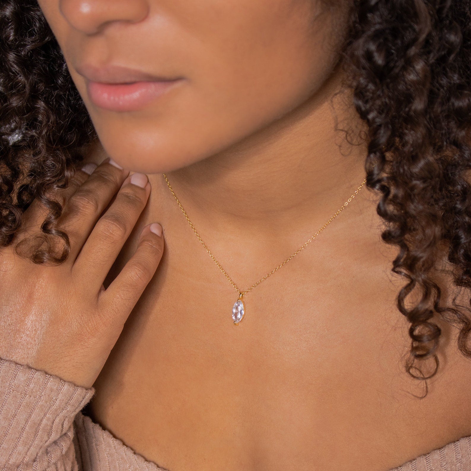A woman wears the Athens Diamond Necklace, featuring a delicate gold cable chain and small oval pendant, touching her chest in an off-shoulder top.