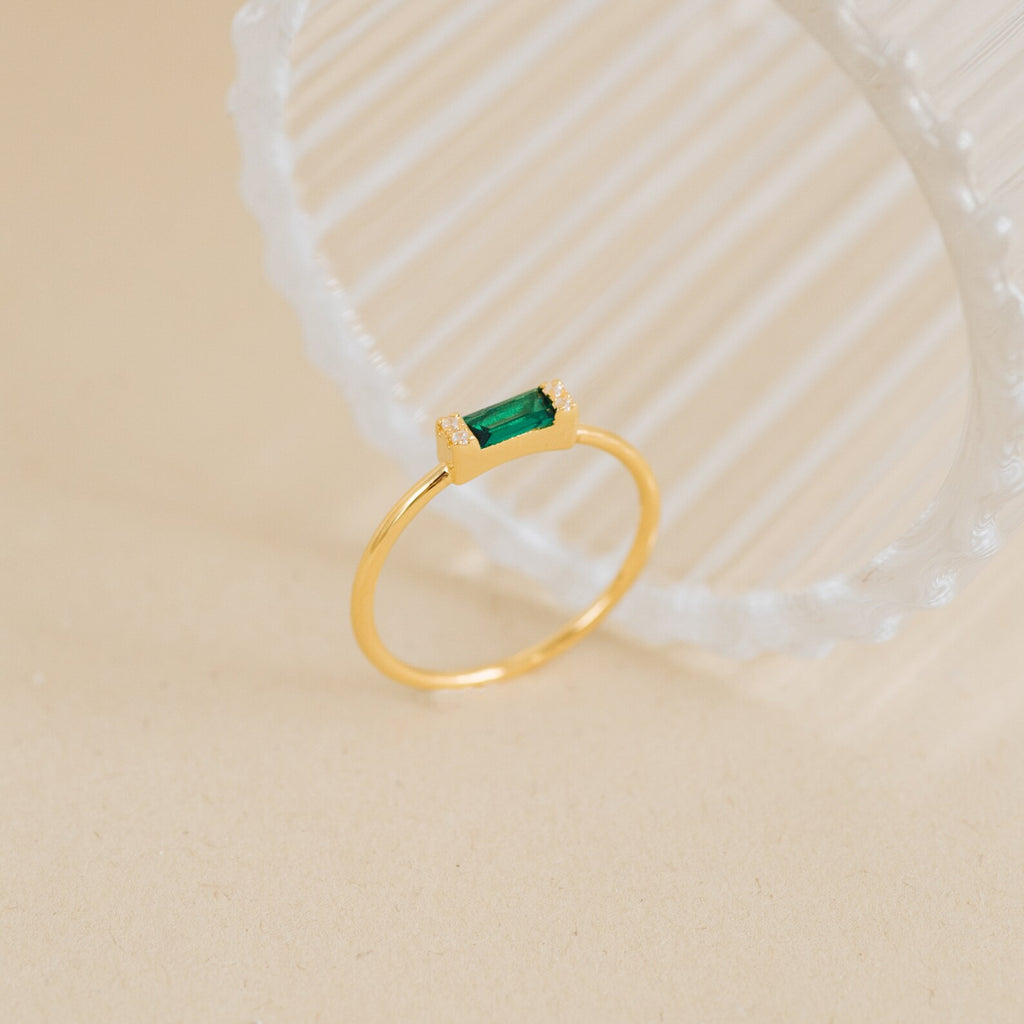 The Empire Emerald Ring, featuring a rectangular green gemstone set on a gold band, is displayed on a light beige surface beside a ribbed glass object.