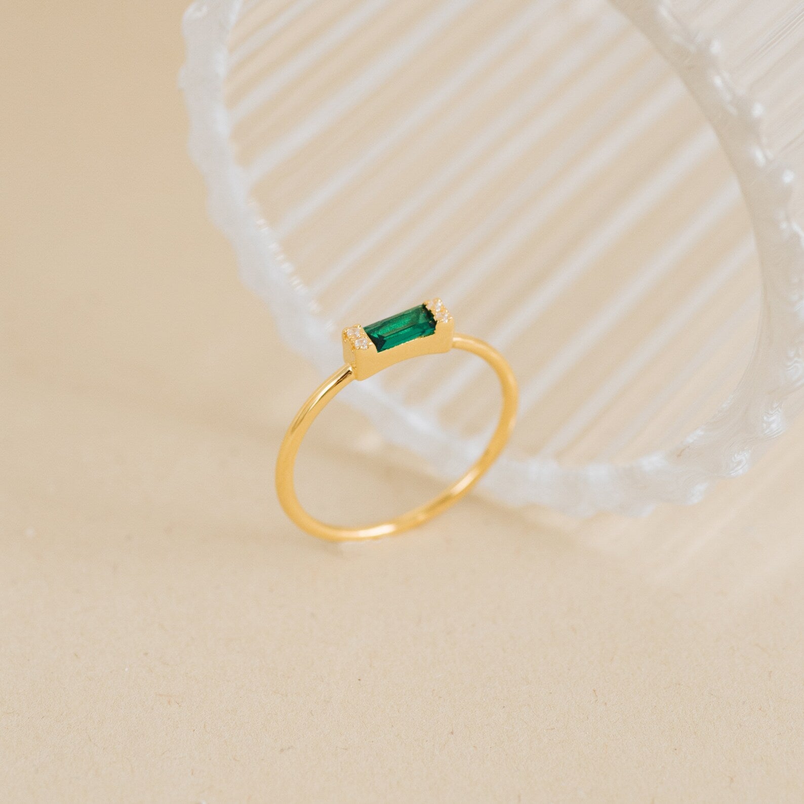 The Empire Emerald Ring, featuring a rectangular green gemstone set on a gold band, is displayed on a light beige surface beside a ribbed glass object.