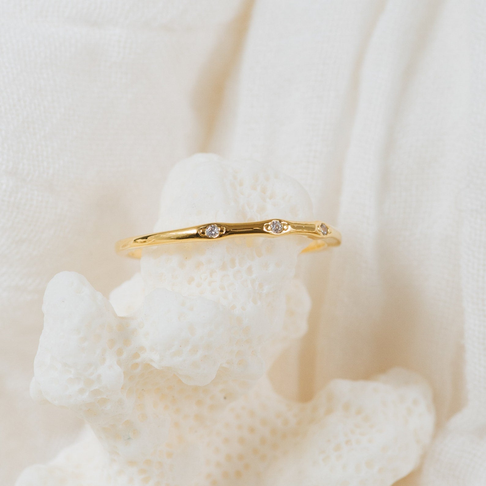 The Juliet Diamond Ring, adorned with three small diamonds, is displayed on white coral against a soft fabric backdrop—an elegant and minimalist piece for jewelry lovers.