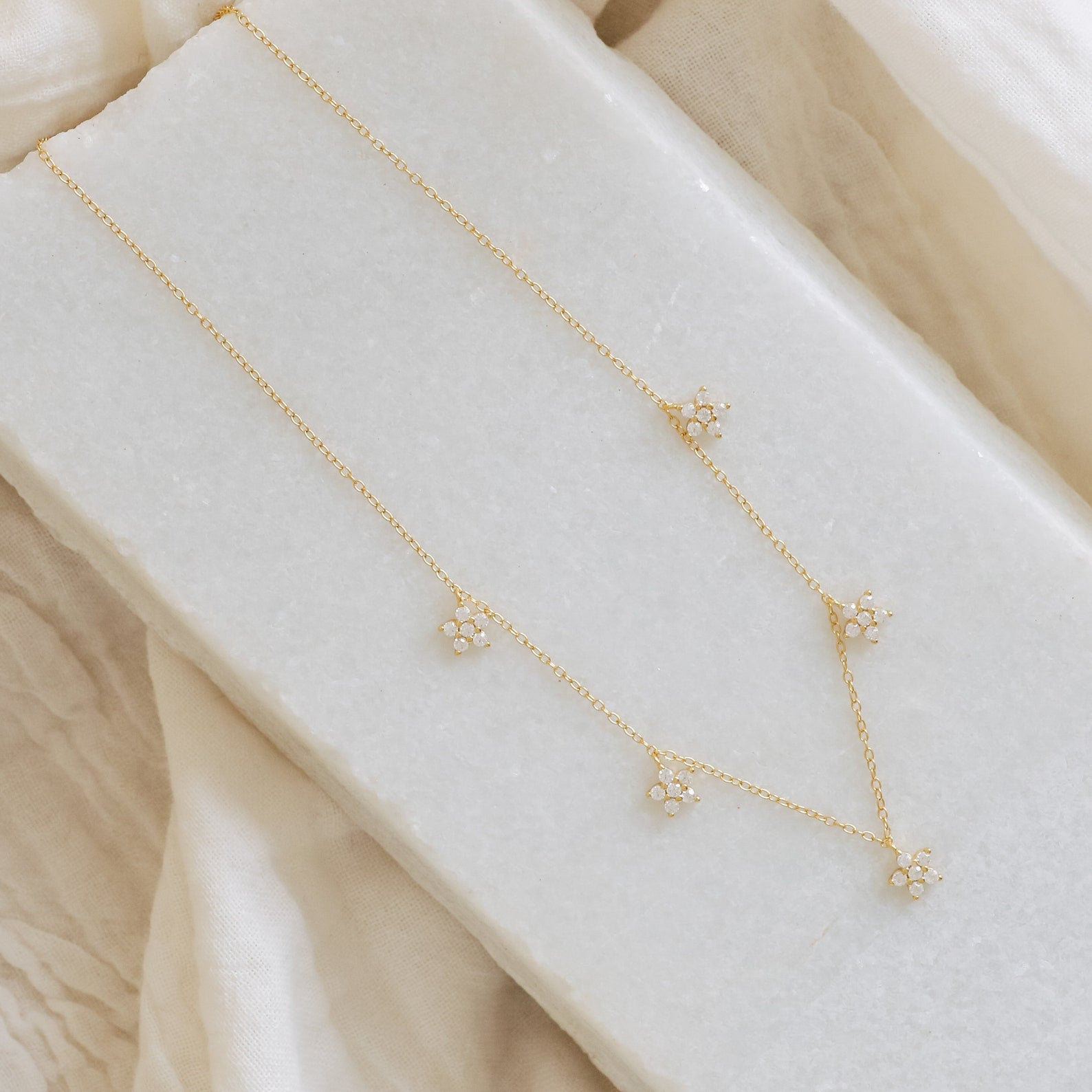The Nanala Diamond Flower Necklace showcases five delicate, diamond-studded flower pendants, presented on a white textured surface with a fabric backdrop.