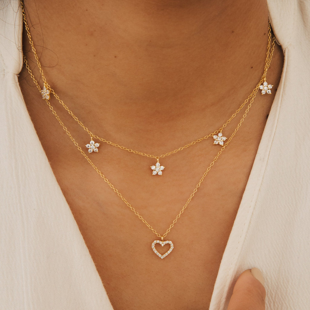 Wearing two gold necklaces: one adorned with crystal stars and the other, the Nanala Diamond Flower Necklace featuring a crystal heart pendant, both styled over a white top.