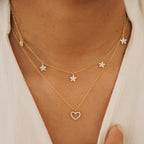 Wearing two gold necklaces: one adorned with crystal stars and the other, the Nanala Diamond Flower Necklace featuring a crystal heart pendant, both styled over a white top.