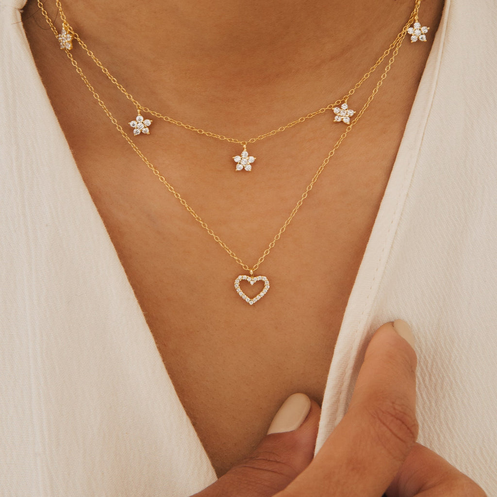 Close-up of the Forever Pave Heart Necklace, a layered gold piece with a sparkling pave heart pendant, worn with a flower necklace on a cream top—an elegant bridal jewelry choice.