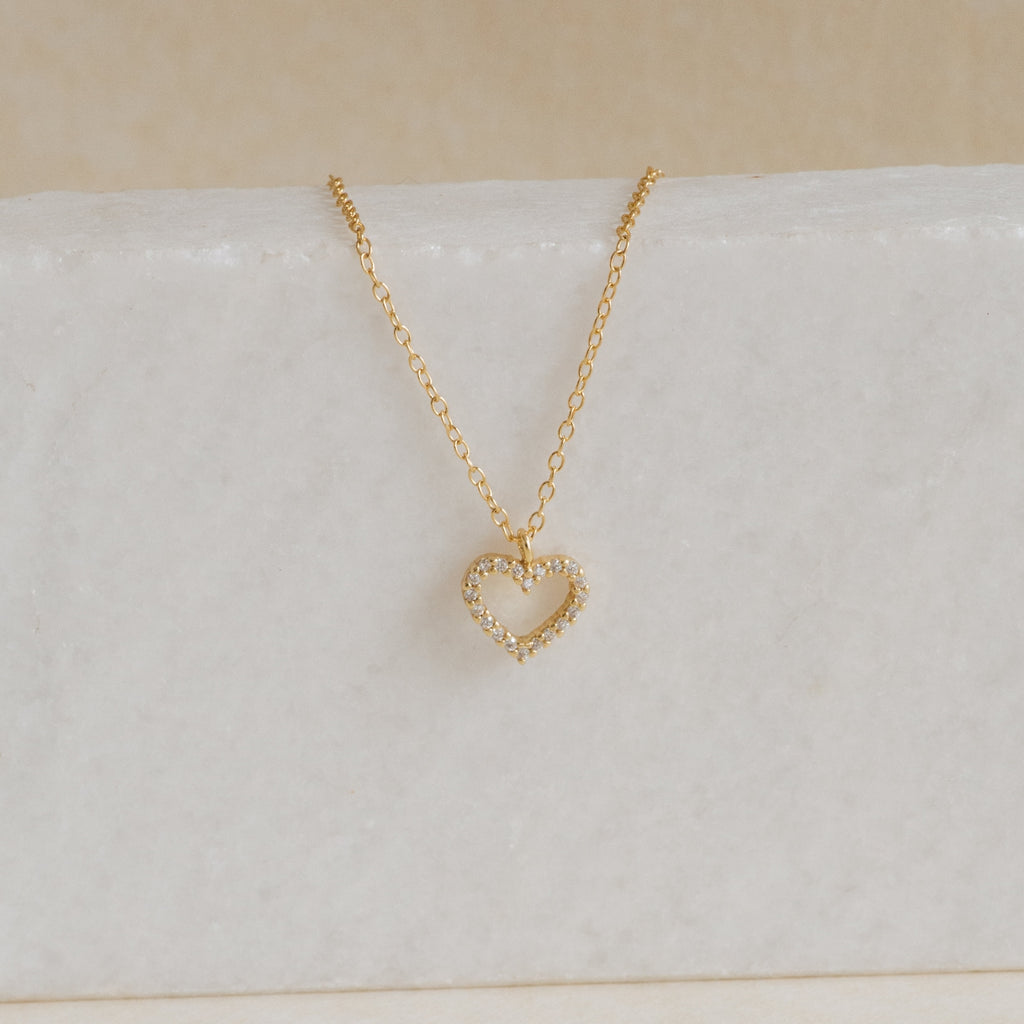 The Forever Pave Heart Necklace features a gold, diamond-studded open heart pendant, elegantly displayed on white marble—perfect for bridal jewelry or special occasions.