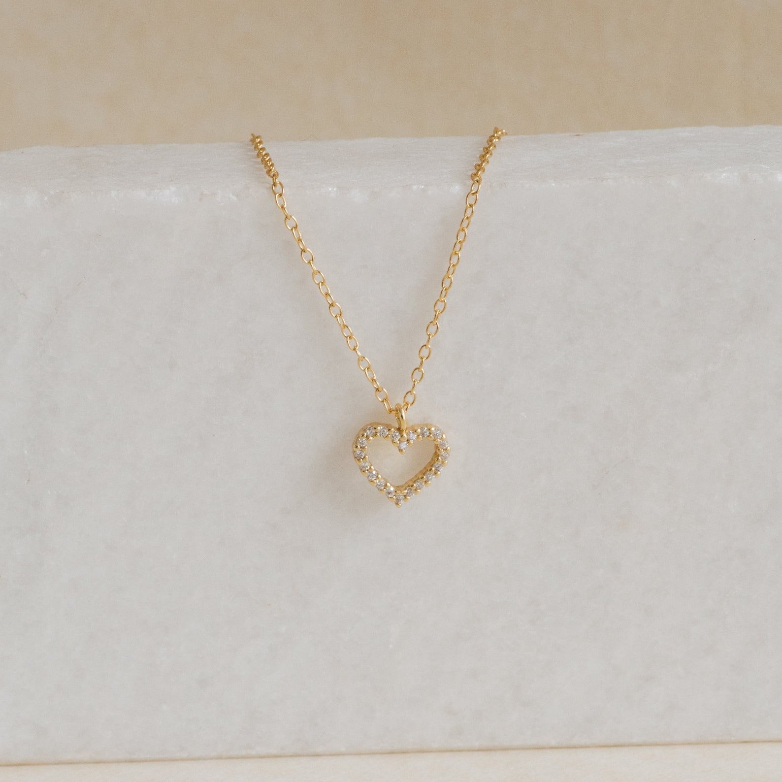 The Forever Pave Heart Necklace features a gold, diamond-studded open heart pendant, elegantly displayed on white marble—perfect for bridal jewelry or special occasions.