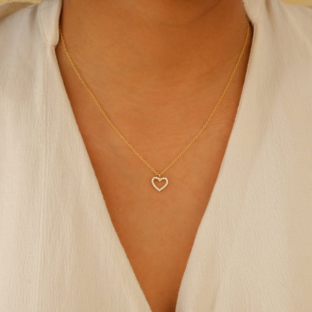 A person wearing the Forever Pave Heart Necklace, featuring a small, shimmering gold heart pendant, over a cream-colored top.