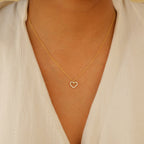 A person wearing the Forever Pave Heart Necklace, featuring a small, shimmering gold heart pendant, over a cream-colored top.