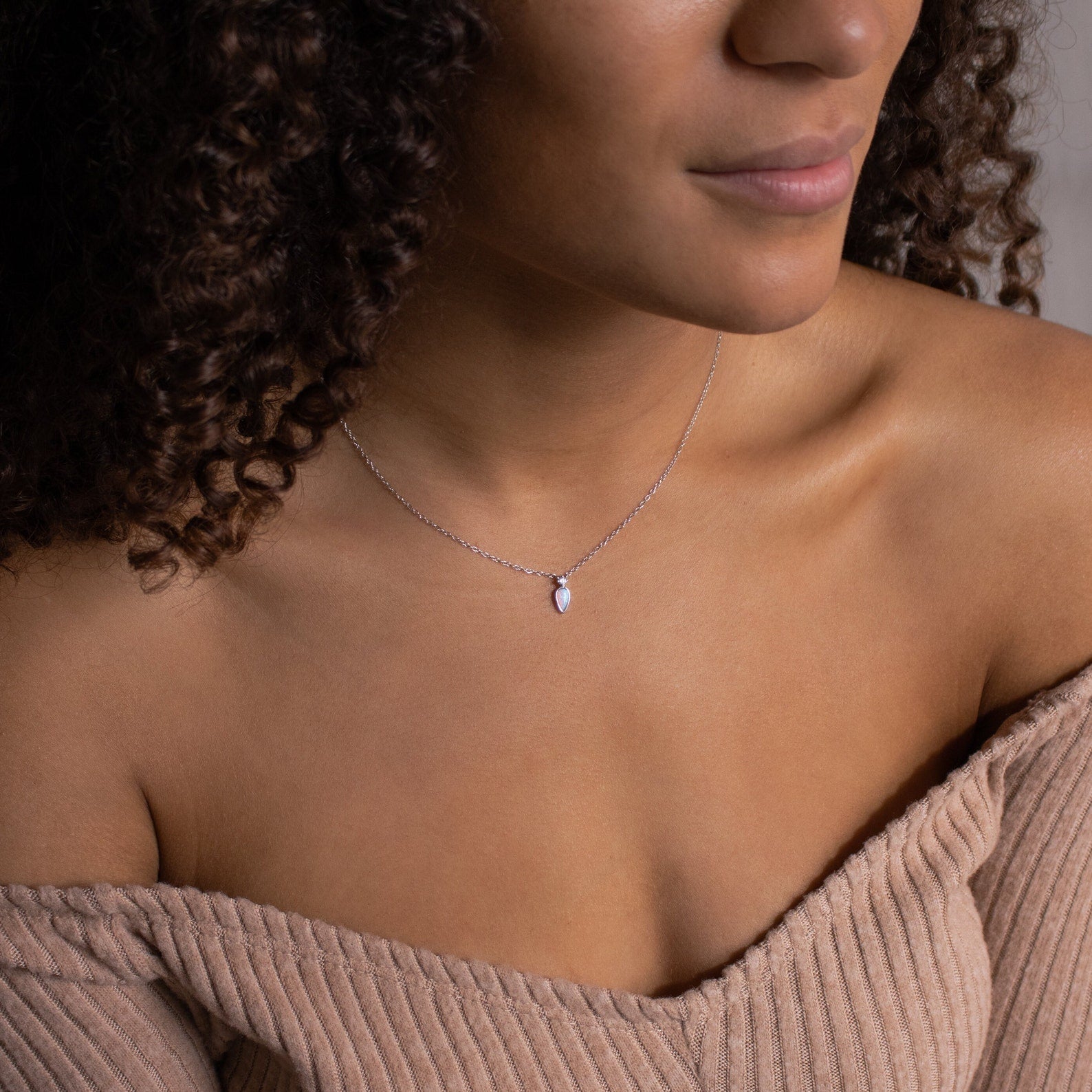 A woman with curly hair wears an off-shoulder ribbed top and the Opal Drop Necklace, featuring a small iridescent opal pendant.