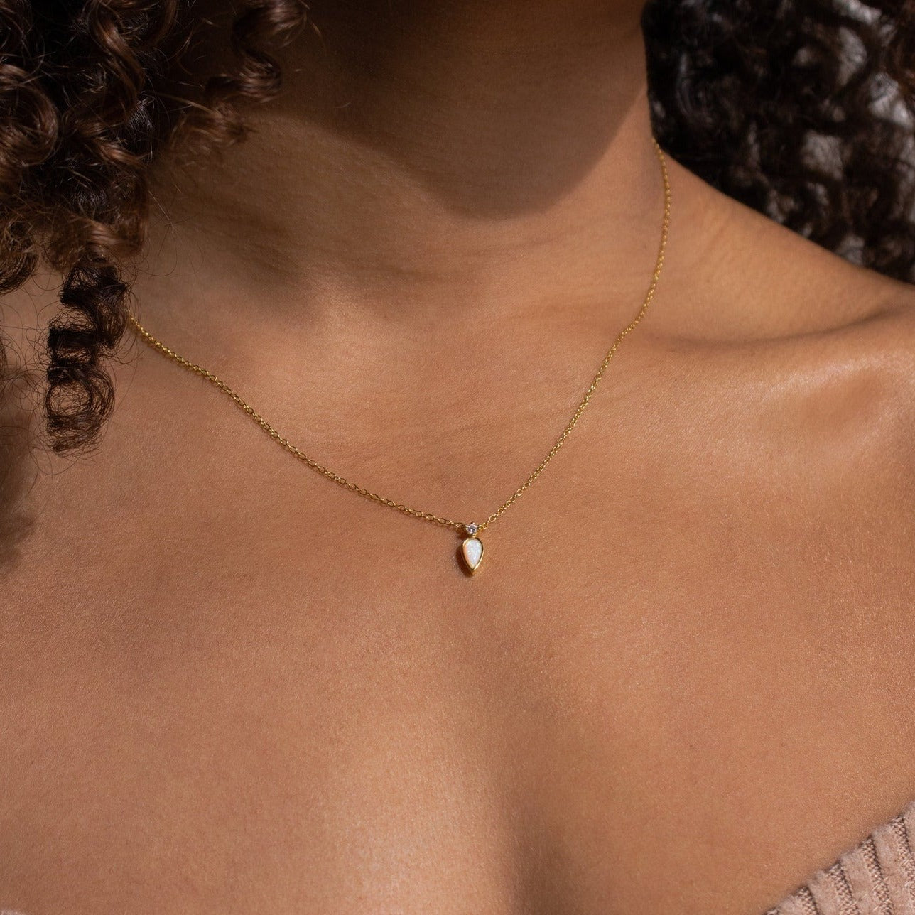 Close-up of a gold necklace with a pear-shaped opal pendant and tiny crystal accent above, worn against bare skin and styled with a taupe ribbed off-shoulder top.