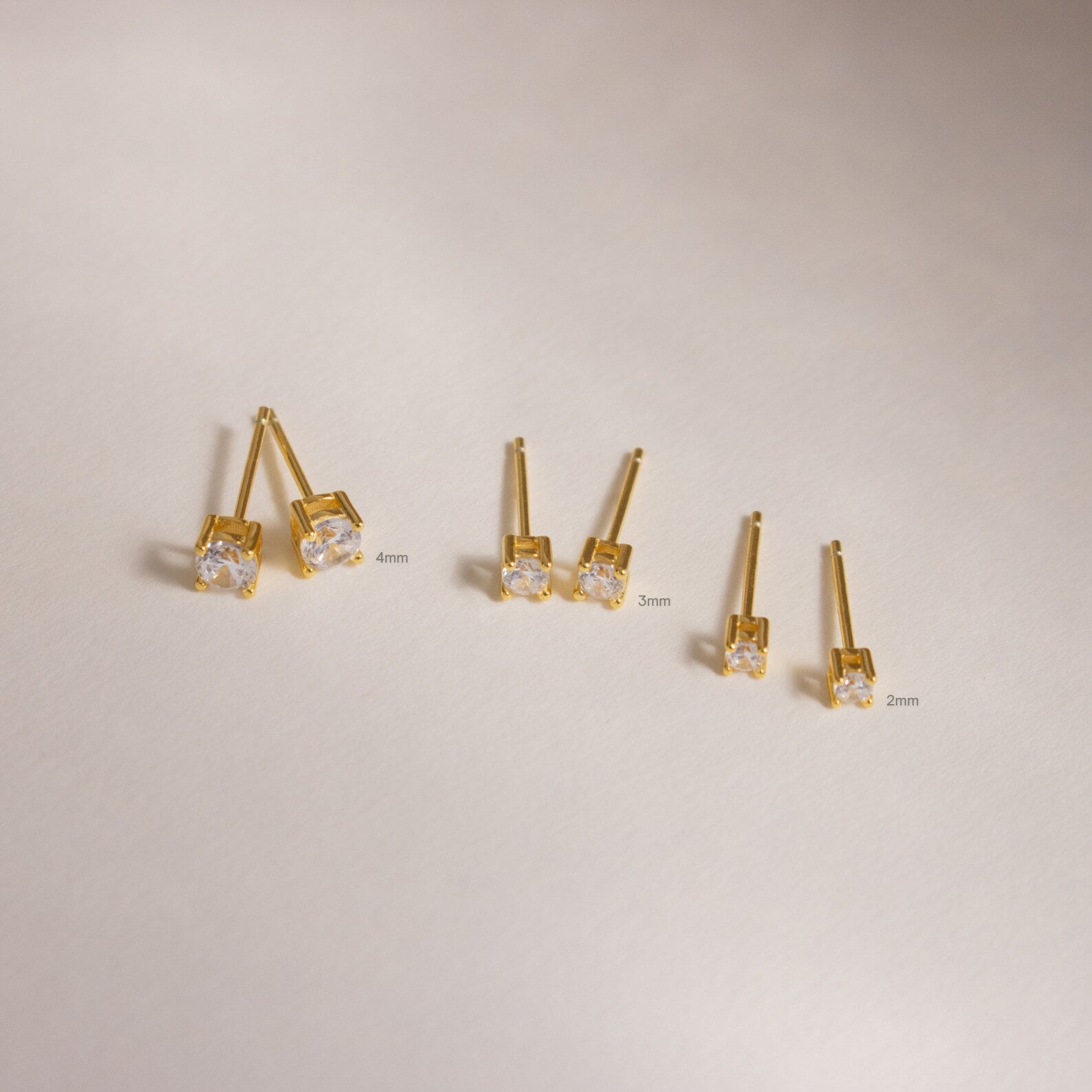 Three pairs of Booker Tiny Diamond Studs in 4mm, 3mm, and 2mm sizes rest on a neutral background—perfect minimalist gold earrings for everyday subtle sparkle.