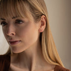 A blonde woman with bangs wears the Booker Tiny Diamond Studs as her everyday earrings, pairing them with a brown top and a thoughtful expression.