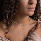 A woman in a beige off-shoulder top touches her shoulder while wearing the Santorini Turquoise Choker adorned with gold and blue gemstones.