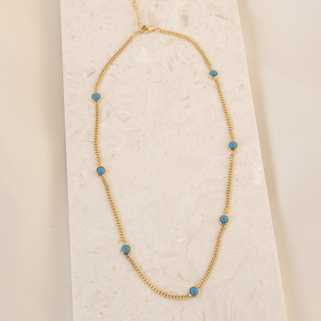 The Santorini Turquoise Choker features a gold chain with turquoise drops, beautifully displayed on a light textured surface.