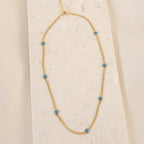 The Santorini Turquoise Choker features a gold chain with turquoise drops, beautifully displayed on a light textured surface.
